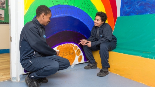 Two students sitting in front of a rainbow painted on a wall that says equality 
