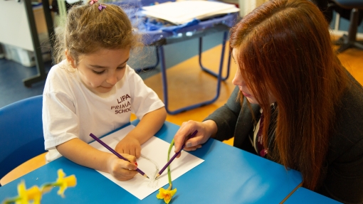 A female pupil sat at a desk with daffodils next to her which she is drawing. A teacher is sat next to her, looking at her drawing