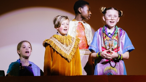Creative cross-curricular learning at River Beach Primary School. A group of 4 children stood on a stage, wearing colourful costumes with a spotlight pointing at them. 