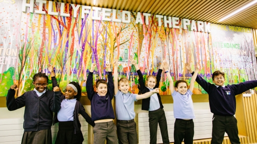 A group of children lined up in front of a large painting on the wall. They are smiling and throwing their arms up in the air. 
