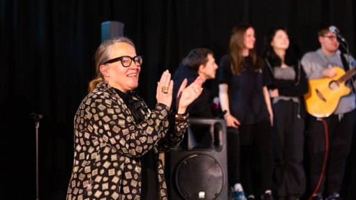 A teacher stands at the forefront of the frame clapping her hands and smiling. In the background are a group of students in a band, holding instruments.