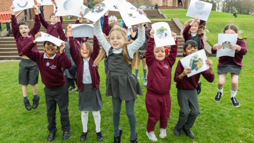 A group of children outside, jumping on the grass, holding up their art work.