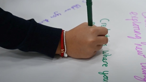 Close-up of a child writing with a green marker on white paper, wearing beaded bracelets. Colorful handwritten words like 'Creating' and 'exploring new things' are visible.