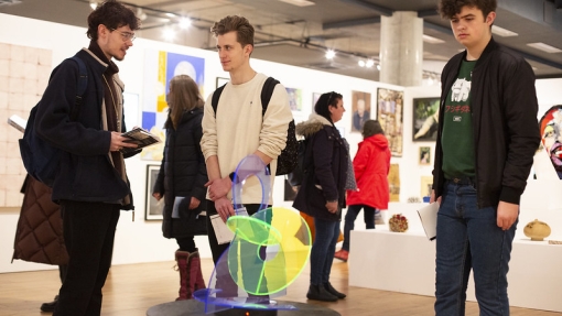 A group of people are observing an art exhibit in a gallery. The exhibit features various artworks displayed on the walls and a colorful, abstract sculpture on a pedestal in the centre. The gallery has wooden floors and bright lighting.