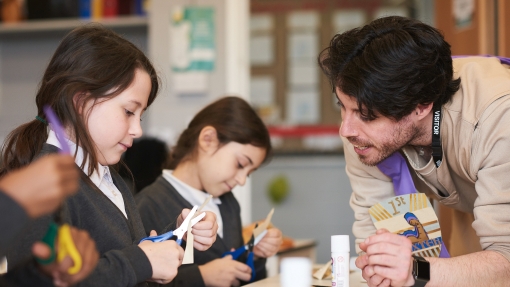 A teacher is leaning on a desk to help two students who are doing a craft activity