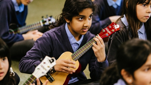 Girl sitting on the floor of a classroom playing a ukulele.