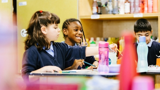 Two young people sitting at a classroom desk with paint materials in the foreground.
