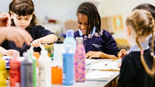 A group of children at a classroom art table. There are colourful bottles of paint in the middle of the table. 