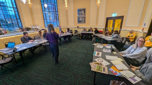 A group of people seated around tables in a meeting room, listening to a person standing and speaking at the front. The room has large windows, framed pictures on the walls, and patterned green carpet.