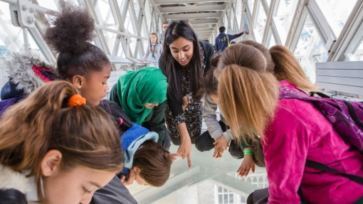 Group of children and an adult leaning over a glass floor on a high-level walkway inside Tower Bridge, looking down at the view below.