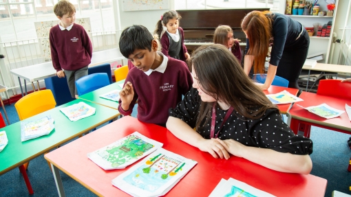 Students looking closely at drawings and designs on the table with a teacher sat on the table at the front and a teacher leaning over a table in the back righthand corner 