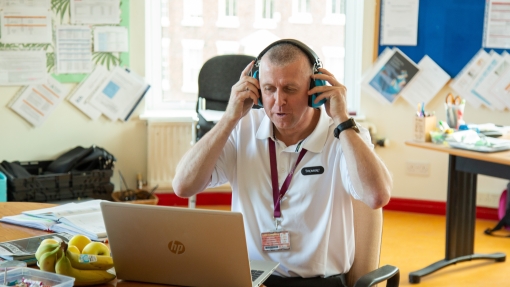 A white male teacher in a white t-shirt sitting in front of a laptop putting ear defenders on