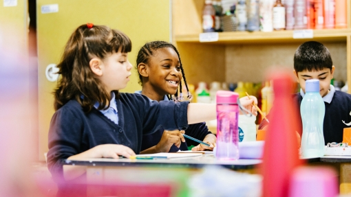 Three school children sit on a table, laughing as they concentrate on painting. 