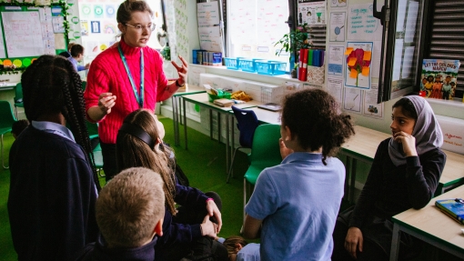 A group pf children sit around in a circle, as their teacher stands with them. They look deep in conversation. 