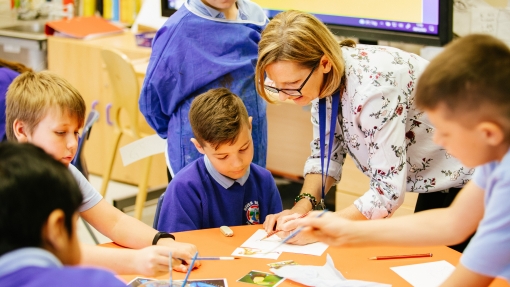 An image of a group of young children sat around a circular table as their teacher demonstrates how to paint something on one of their pieces of paper. 