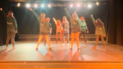 A group of young people standing on a small stage with their backs to the camera and their left hand pointing upwards