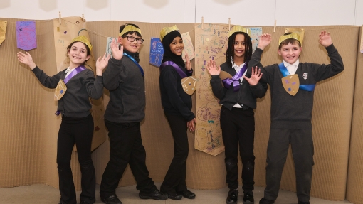 Five students standing in front of art waving wearing crowns