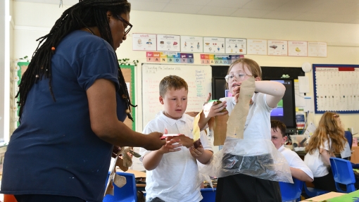 Teacher interacting with two children in a classroom, helping them with a craft activity using paper and other materials. The classroom has blue chairs, colourful displays on the walls, and a large table covered with art supplies.
