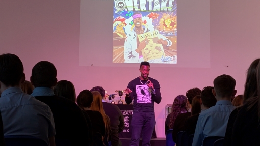 An artist stands in front of an audience giving a music workshop, with a colorful illustrated poster titled “OVERTAKE” projected on the wall behind. The poster includes dynamic cartoon-style graphics and the text “@ShaoDowMusic” at the top. The room is filled with seated students facing the presenter.