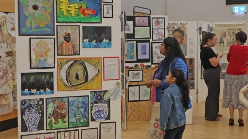 Art exhibition in a school hall displaying colorful student artwork on panels, including paintings, drawings, and mixed media pieces, with visitors observing the displays.