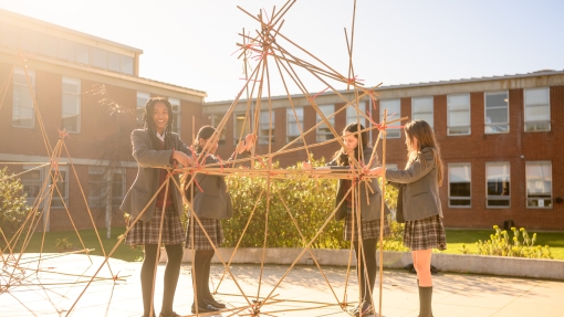 Students in school uniforms working together outdoors to build a large geometric structure made of bamboo sticks and connectors, with a school building in the background.