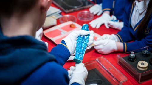 Close-up of a child wearing white gloves holding a bright blue ancient-style figurine, with other small artifacts and display trays on a red table.