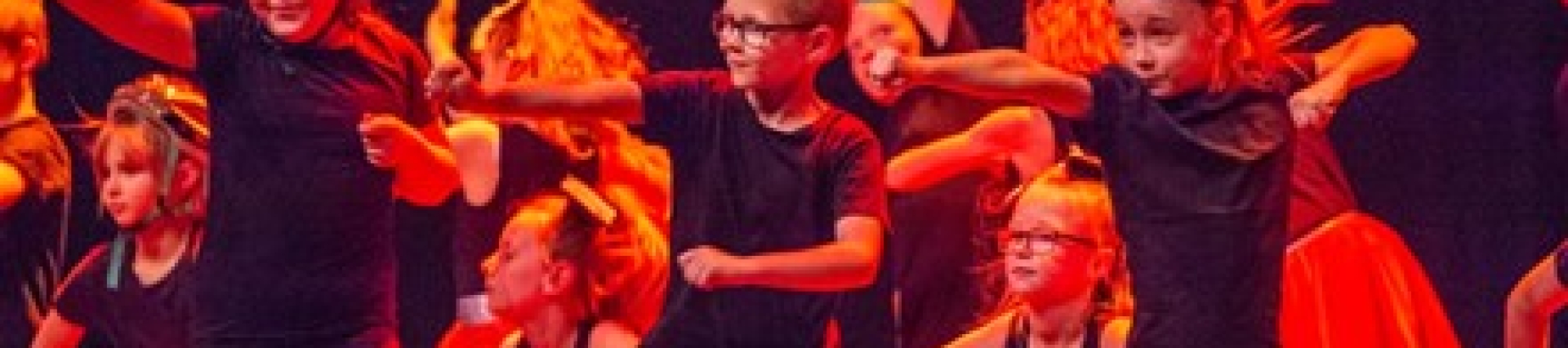 A group of children dance on a stage in dark red lighting. 