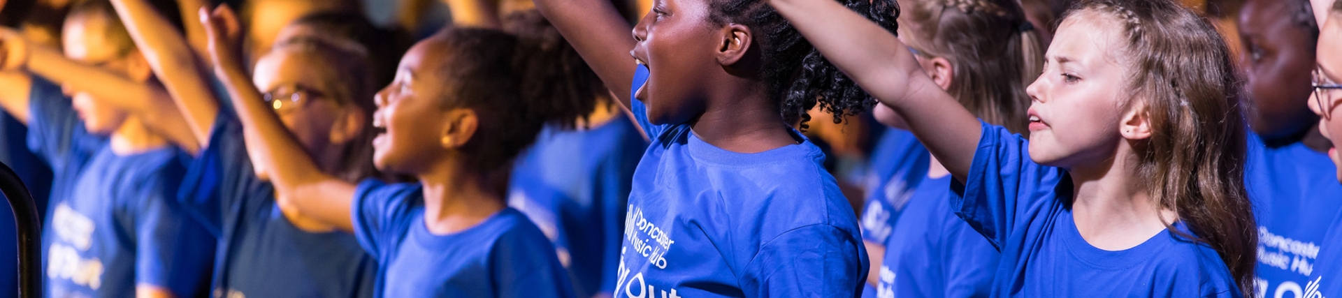 A group of primary aged children in blue teachers singing and raising one arm in the air