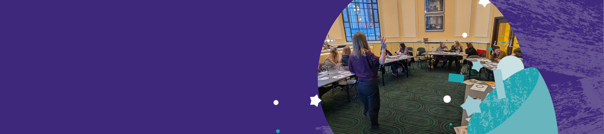 A group of people seated around tables in a meeting room, listening to a person standing and speaking at the front. The room has large windows, framed pictures on the walls, and patterned green carpet.