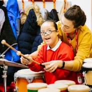 Young primary pupil being taught to play bongos by her teacher who is standing behind her