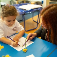 A female pupil sat at a desk with daffodils next to her which she is drawing. A teacher is sat next to her, looking at her drawing