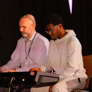 A teacher and a student are sitting together, playing piano.