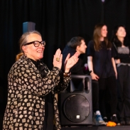 A teacher stands at the forefront of the frame clapping her hands and smiling. In the background are a group of students in a band, holding instruments.