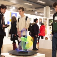 A group of people are observing an art exhibit in a gallery. The exhibit features various artworks displayed on the walls and a colorful, abstract sculpture on a pedestal in the centre. The gallery has wooden floors and bright lighting.