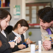 A teacher is leaning on a desk to help two students who are doing a craft activity