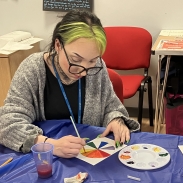 Person with green and black hair paints a colour wheel at a table with art supplies