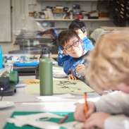 A student wearing glasses and a blue overall is painting and smiling in a classroom