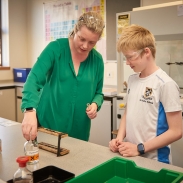 A teacher in a green top is helping a student in a white t shirt do a science experiment