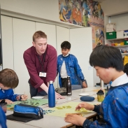 A teacher in a burgundy shirt leans in to help a group of students sitting at a table with their painting.