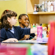 Two young people sitting at a classroom desk with paint materials in the foreground.