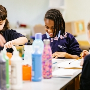 A group of children at a classroom art table. There are colourful bottles of paint in the middle of the table. 