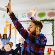 A teacher at the front of a classroom with his arms raised in the air. There are children in the background with their hands also in the air. 