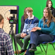 People seated in front of a green screen being filmed by a camera on a tripod.