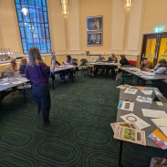 A group of people seated around tables in a meeting room, listening to a person standing and speaking at the front. The room has large windows, framed pictures on the walls, and patterned green carpet.
