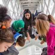 Group of children and an adult leaning over a glass floor on a high-level walkway inside Tower Bridge, looking down at the view below.